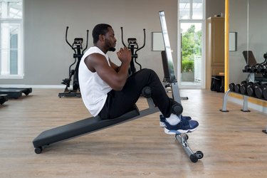 a young adult wearing a white t-shirt and black pants uses a sit-up bench at the gym